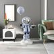 The Cosmic Sky Astronaut Butler Balloon Sculpture by Giant Sculptures stands 110cm tall in a light blue spacesuit with a reflective helmet, holding a moon-shaped balloon. It beautifully complements the decor, including the modern rooms green sofa and white cabinet with flowers.