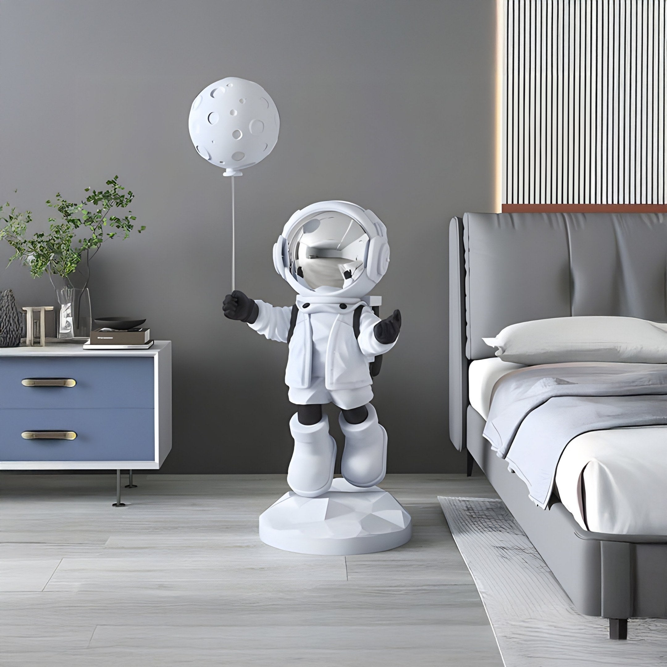 A Nova White Astronaut Moon Balloon Sculpture by Giant Sculptures stands in a modern bedroom with a blue side table holding plants and books next to a gray bed with white bedding, featuring minimalist decor and a striped wall accent.