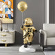 The Celestial Gold Astronaut Ranger Balloon Sculpture by Giant Sculptures, with its shiny gold finish and celestial balloon, stands on a white base. It complements modern decor alongside abstract art, a white sideboard, and an inviting white sofa.