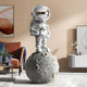 A Giant Sculptures Lunar Silver Astronaut Star On Moon Sculpture (125cm) brings interplanetary flair to a modern living space with a brown couch, abstract art, and a white console table decorated with plants and décor elements against beige walls.