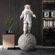 A Moonwalk-Inspired Display showcases the 128cm White & Grey Space Astronaut Moonwalk Sculpture by Giant Sculptures, set on a lunar surface. The modern room features dark walls, a small pink cabinet with a plant, and a bookshelf filled with space-themed décor.