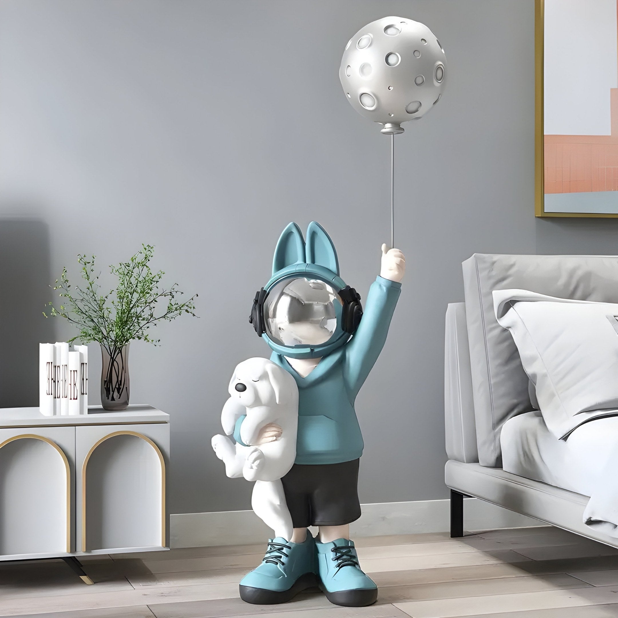 The Blue Astronaut Bunny Pup Balloon Sculpture by Giant Sculptures is a whimsical 130cm statue featuring rabbit ears, a moon-shaped balloon, and a small animal. It comes with headphones and stands on the floor in a modern room with cosmic whimsy, complete with bed and side table.