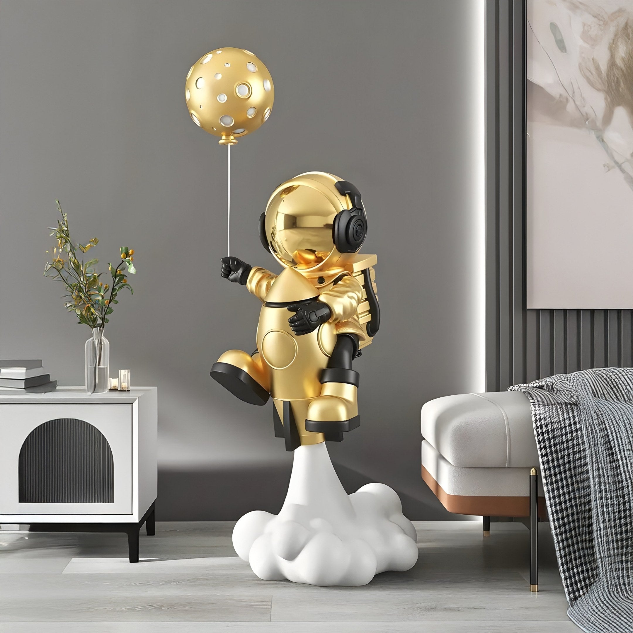 In a modern room, the Celestial Gold Astronaut Rocket Lift-Off Balloon Sculpture by Giant Sculptures stands at 130cm on a cloud base. The glossy gold astronaut wears headphones and clutches a moon balloon, with a white cabinet and gray bed in the minimalist, elegant backdrop.