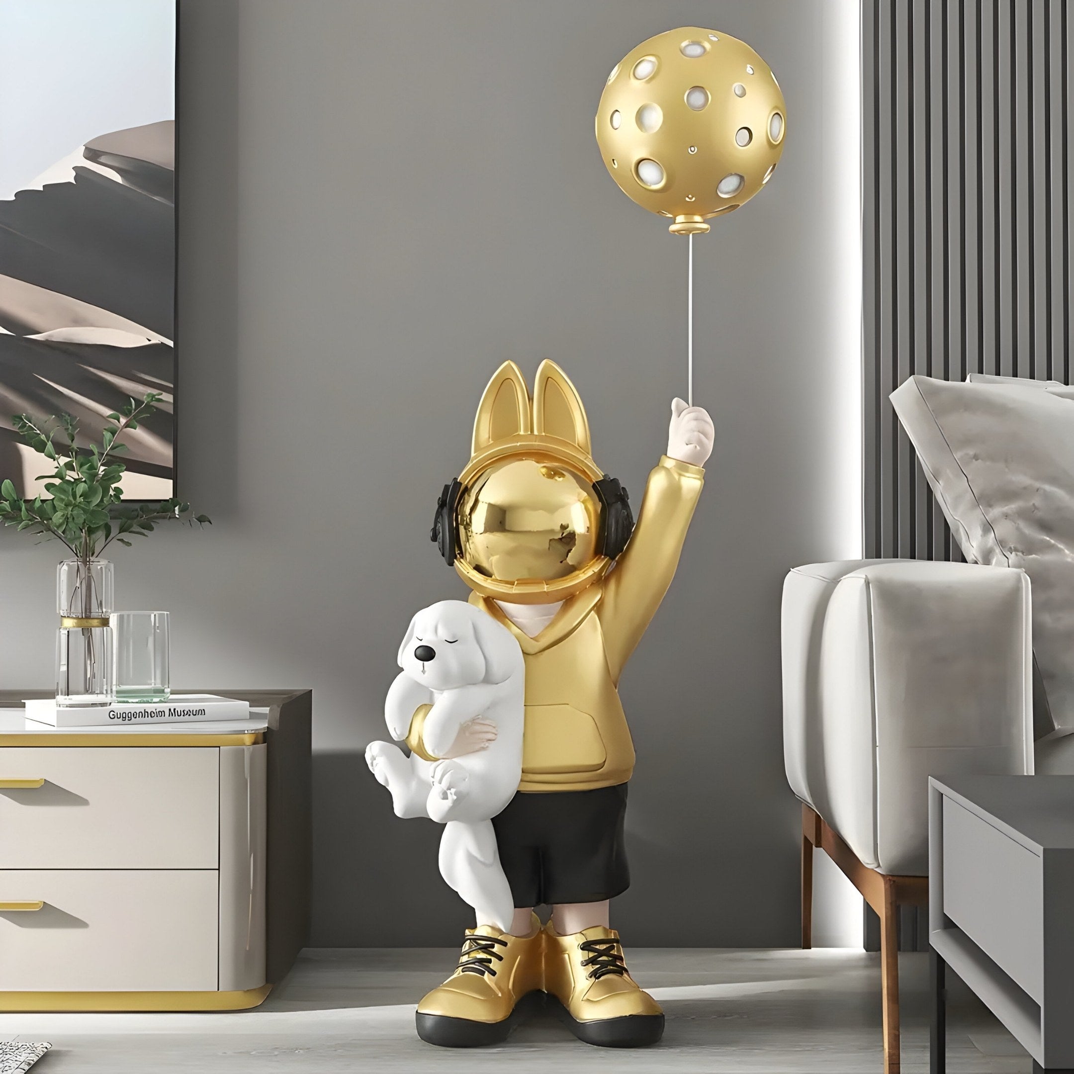 The Gold Astronaut Bunny Pup Balloon Sculpture - 130cm by Giant Sculptures features a golden figure with bunny ears, headphones, and a hoodie holding a dog and polka-dotted balloon. It complements modern rooms with neutral tones and fits perfectly next to plants on cabinets.