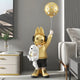 The Gold Astronaut Bunny Pup Balloon Sculpture - 130cm by Giant Sculptures features a golden figure with bunny ears, headphones, and a hoodie holding a dog and polka-dotted balloon. It complements modern rooms with neutral tones and fits perfectly next to plants on cabinets.