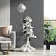 A metallic Lunar Silver Astronaut Rocket Lift-Off Balloon Sculpture from Giant Sculptures stands on a cloud base beside a modern couch and tall green potted plant. Its reflective surface shines against the dark wall, enhancing its space-inspired aura.