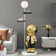 A modern living room includes a white sofa, brown pillow, and black and white lighting, highlighted by the Gold Astronaut Beats Floor Lamp With Shelves by Giant Sculptures. A gray wall and sideboard featuring a vase of dried flowers complete the decor.