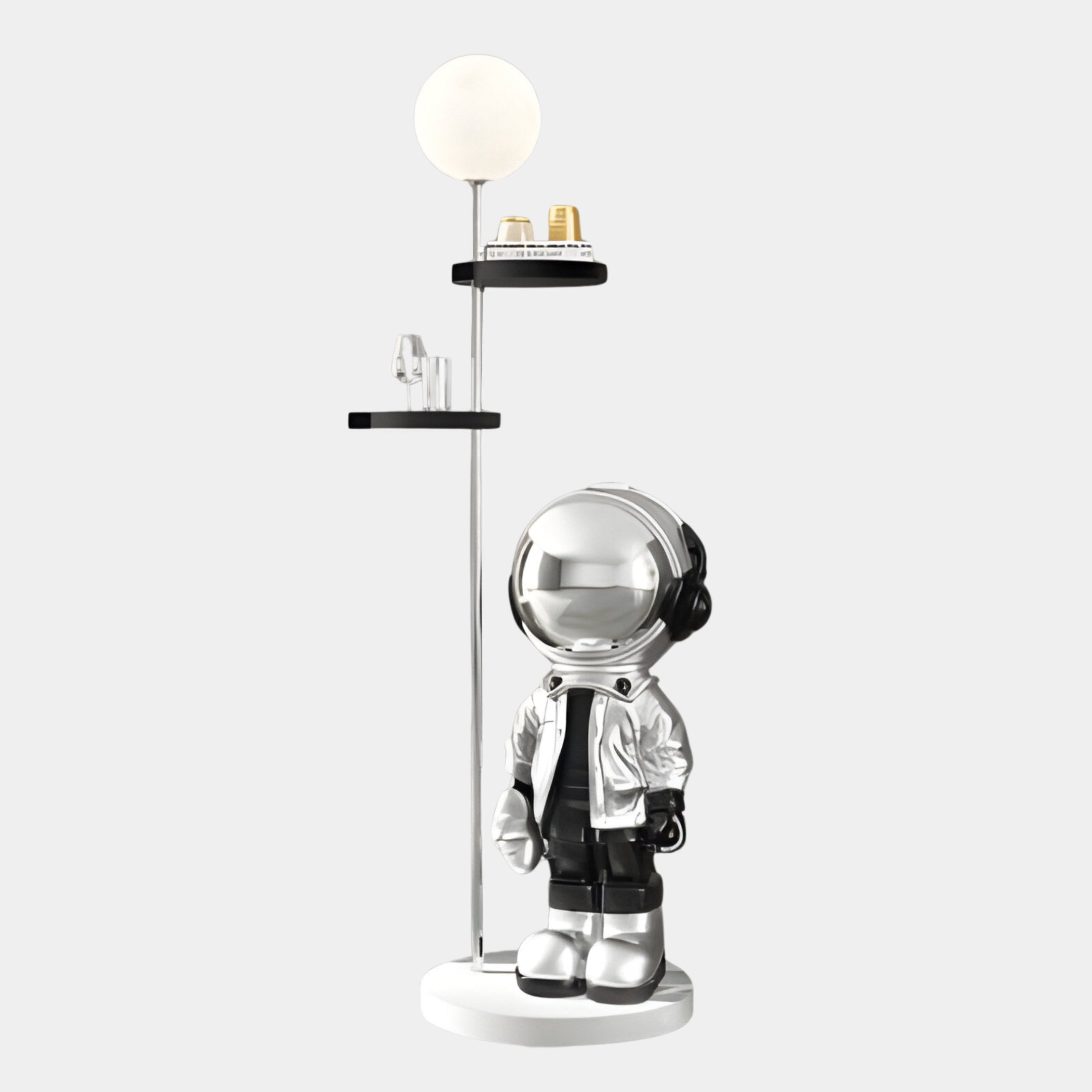 A silver astronaut figure from Giant Sculptures stands beside the Silver Astronaut Beats Floor Lamp With Shelves - 135cm, ideal for holding small items. The robot features a reflective visor and metallic boots, adding a unique space-themed touch against the plain gray background.