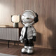 A sleek Lunar Silver Astronaut Beats Sculpture - 150cm by Giant Sculptures adds a futuristic touch to a wooden-walled and floored room. The space-inspired piece, in silver with black accents, including a helmet and jacket, is complemented by a modern lamp on the sideboard for added coziness.