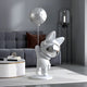 A modern, minimalist room features Giant Sculptures Cosmic Chrome Astronaut Bunny Moon Balloon Sculpture - 150cm. The decor includes a grey sofa, a brown pillow, a round white table with books, and a tall lamp in the background.