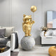 The Gilded Gold Astronaut Kitty Moon Balloon Sculpture by Giant Sculptures, standing at 150cm, graces a modern living room with sleek white and gray furnishings alongside a vase of flowers on the side table.