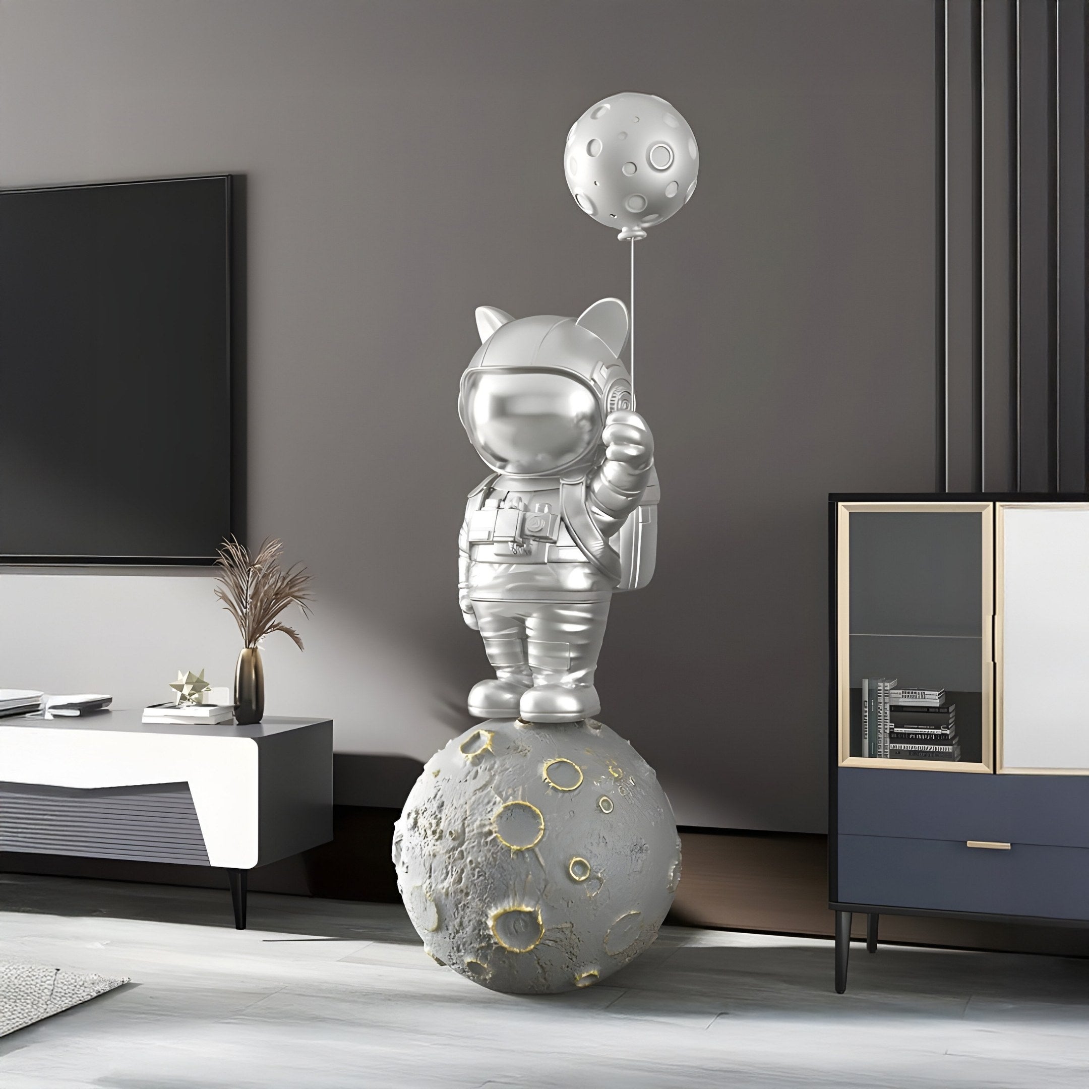 The 150cm Cosmic Chrome Astronaut Kitty Moon Balloon Sculpture by Giant Sculptures stands on a moon-like sphere, holding a planet-shaped balloon in a modern living room with gray walls, a TV, plants, and a cabinet.