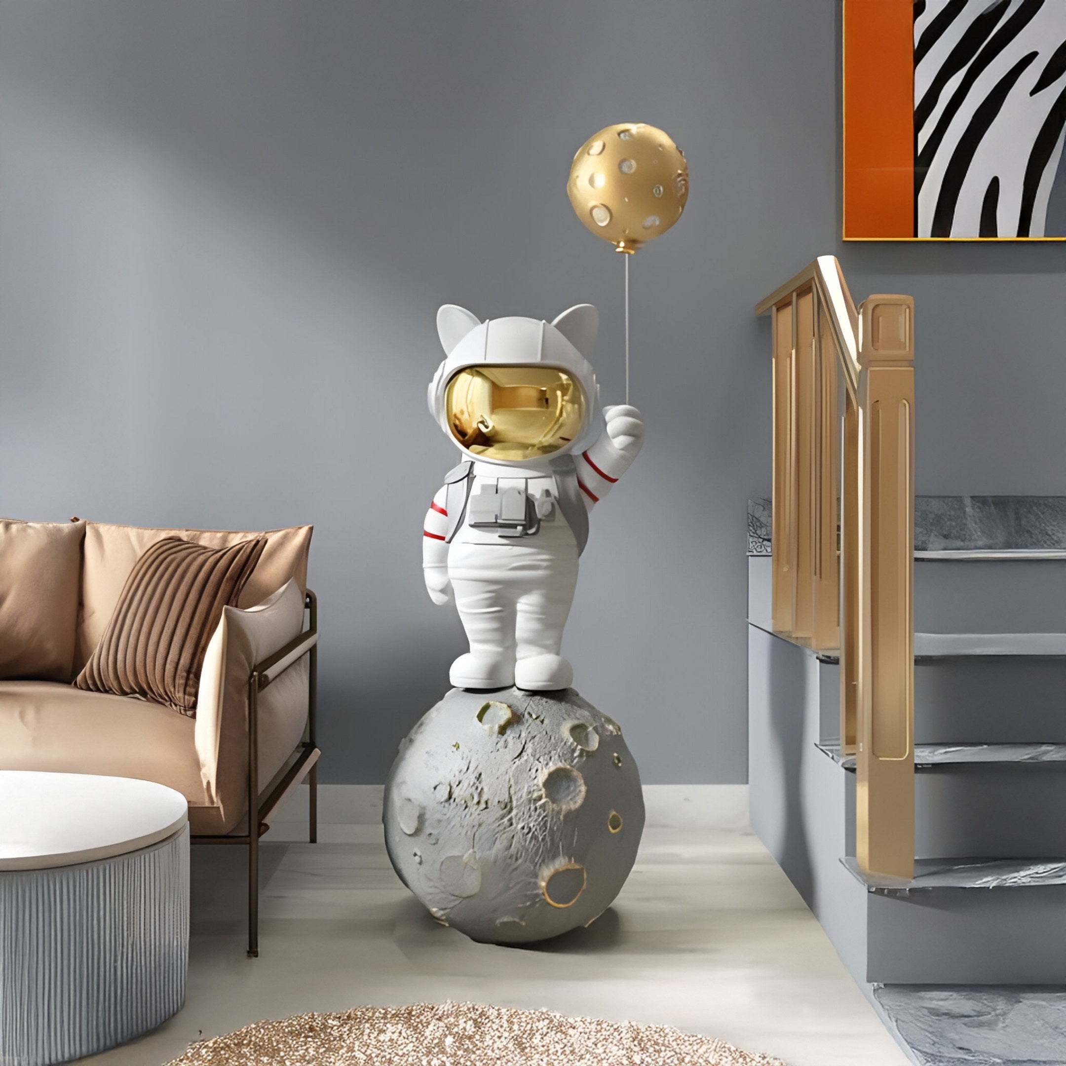 The Nova White Astronaut Kitty Moon Balloon Sculpture by Giant Sculptures, featuring a small astronaut kitty with a gold visor on a moon-like sphere holding a planet-shaped balloon, adds charm to any modern room and complements beige sofas, round white tables, and stairs with gold accents.