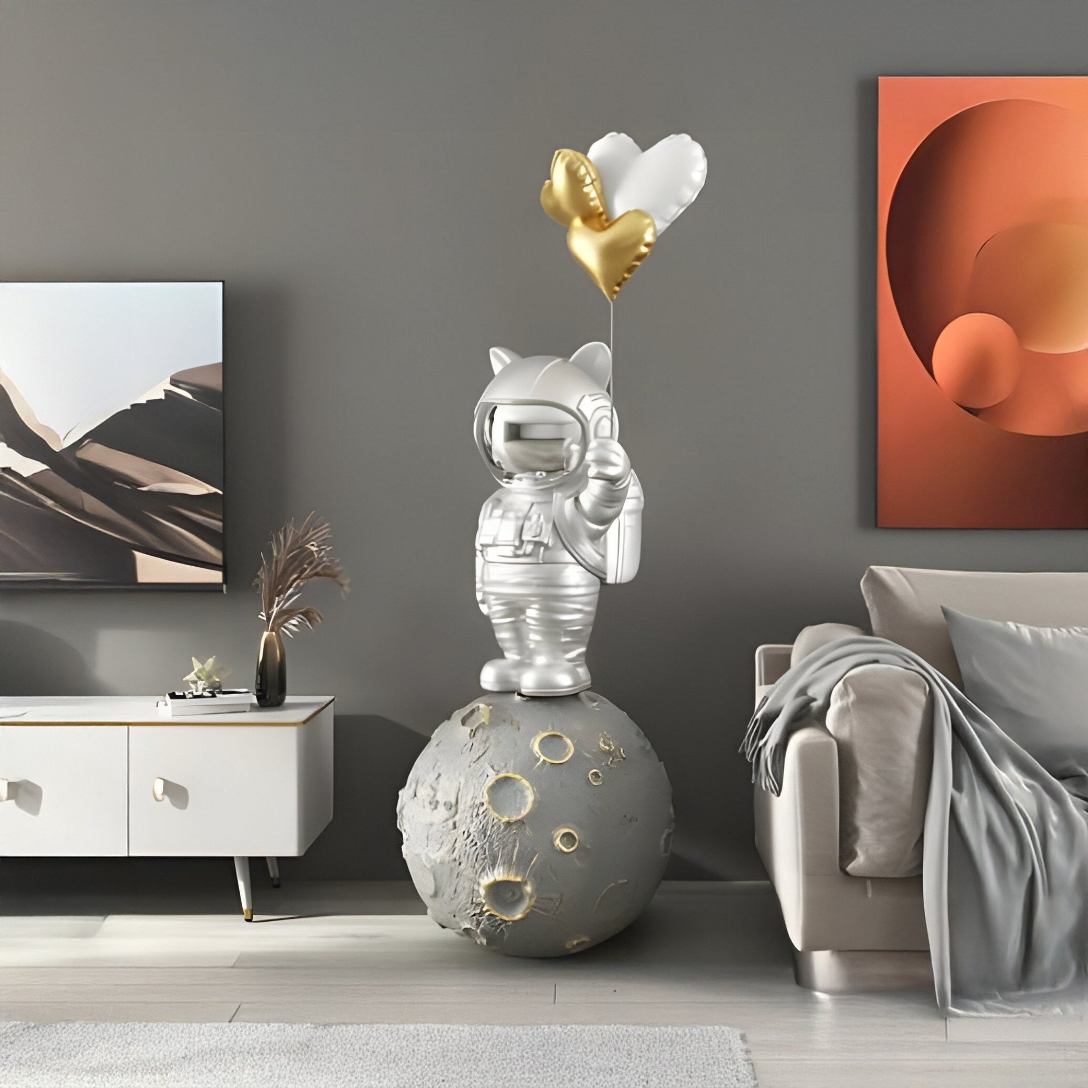 A Cosmic Chrome Astronaut Kitty Heart Balloons Sculpture by Giant Sculptures, standing 160cm tall with heart-shaped silver and gold balloons on a moon-like base, adorns a modern living room with gray walls, abstract art, white console, and beige sofa with a throw blanket.