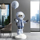 The Cosmic Sky Astronaut Moon Balloon Sculpture by Giant Sculptures is a 160cm resin piece featuring a light blue astronaut with a reflective visor, holding a moon-shaped balloon. It stands in a modern setting with celestial displays, showcasing chairs and large windows that overlook the cityscape.