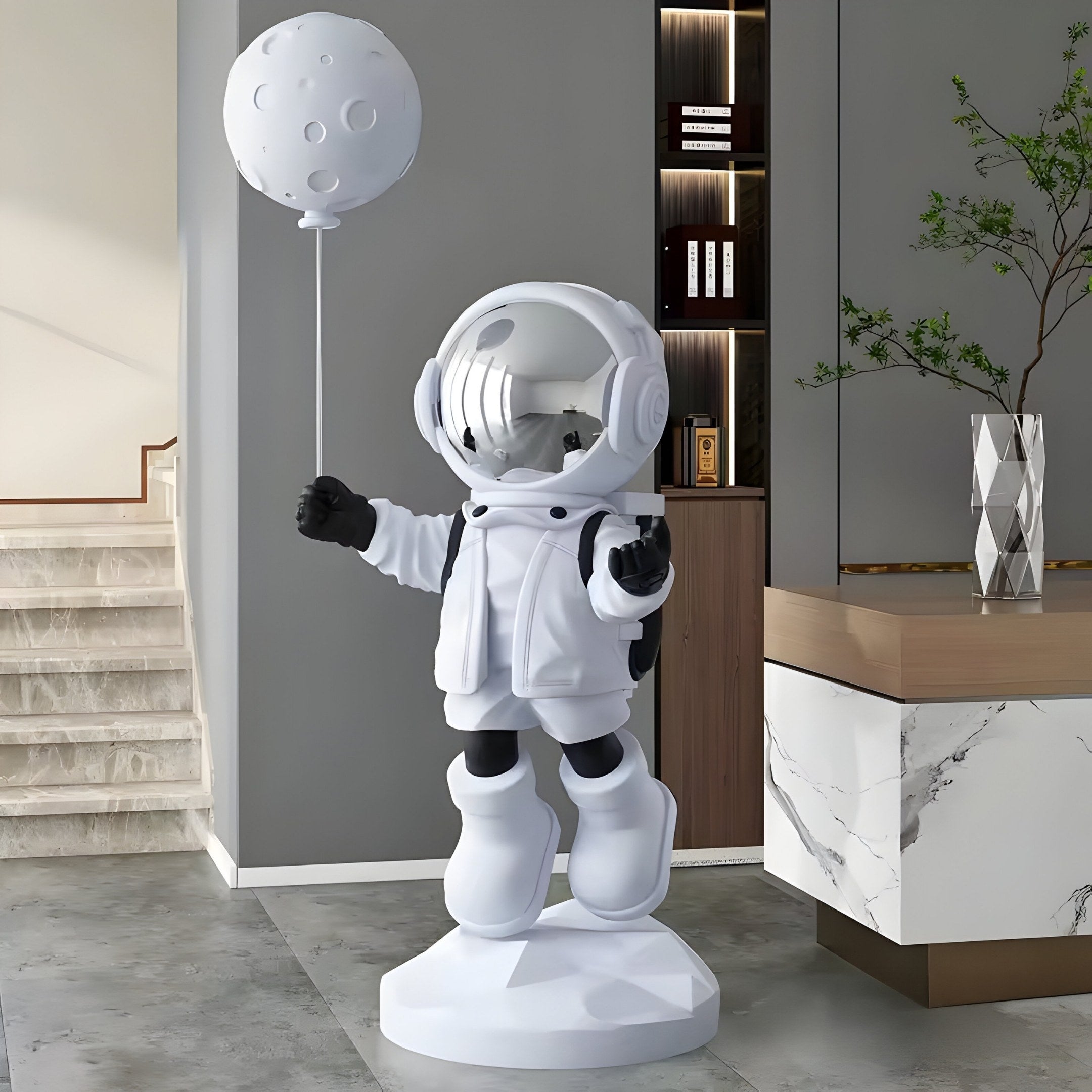 The 160cm Nova White Astronaut Moon Balloon Sculpture by Giant Sculptures stands on a pedestal in a modern room, featuring a reflective visor and puffy boots. Stairs, a plant, and a marble-topped counter complete the background.