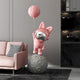 The Candy Pink Astronaut Bunny Moonwalk Balloon Sculpture by Giant Sculptures, standing 180cm tall, whimsically decorates the pink sofa and patterned pillow on a moon-like sphere. This charming piece enhances indoor spaces against a dark textured wall with shelves.