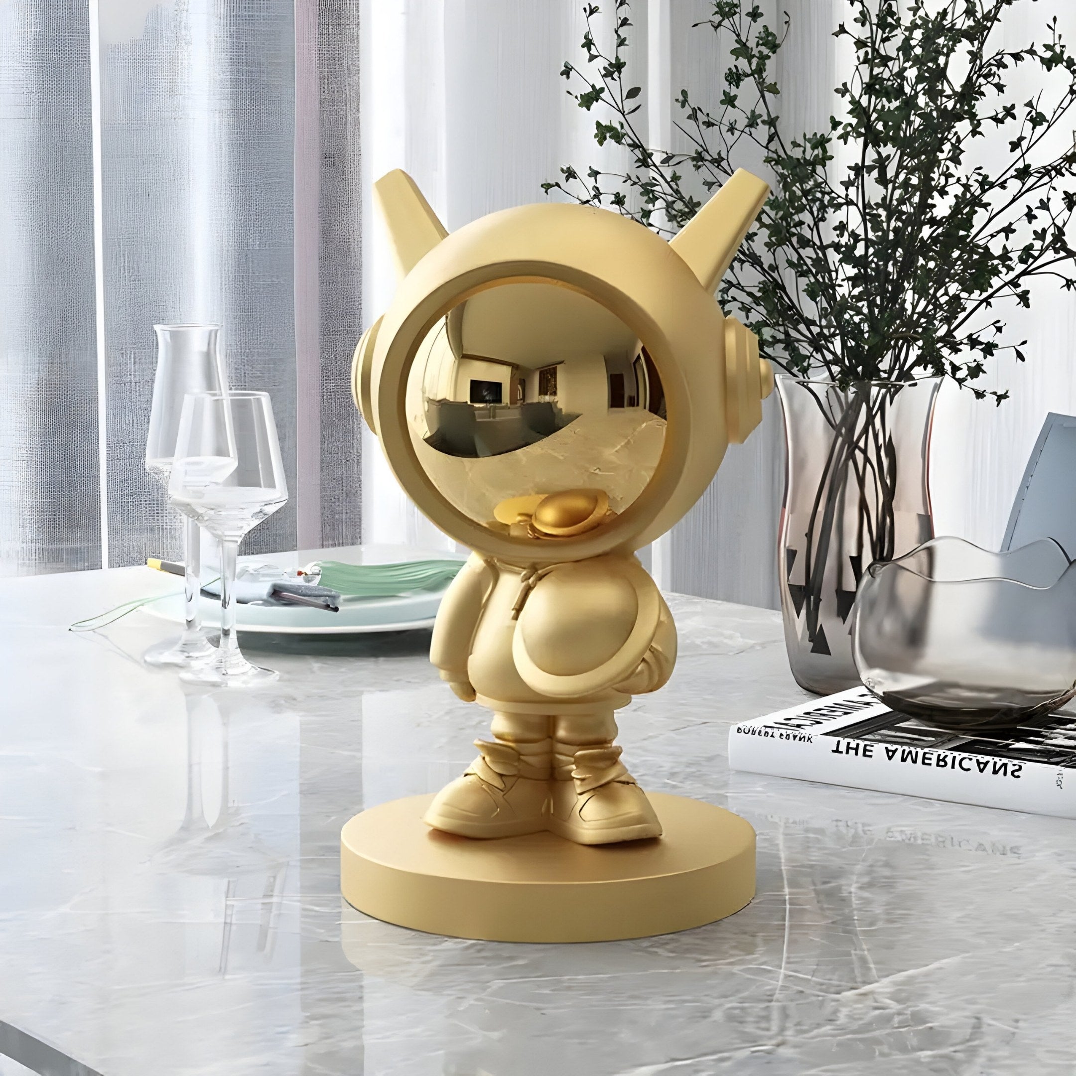 Amidst a cosmic aesthetic, a 30cm Celestial Gold Astronaut DJ Figure from Giant Sculptures stands on glossy marble. Nearby, two elegant glasses sit with a plant and books, while light filters through sheer curtains, casting an ethereal glow.