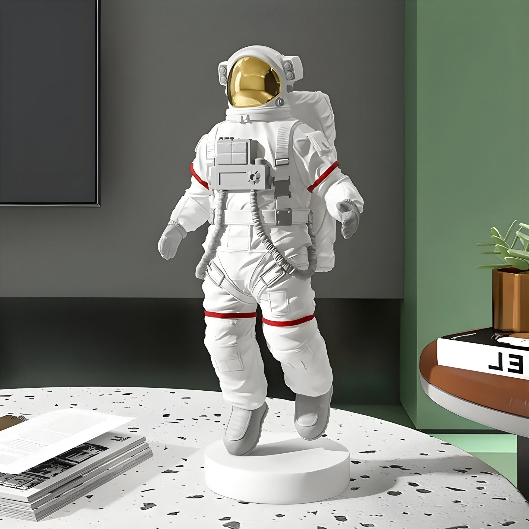 The 30cm White Space Astronaut Voyager Figure by Giant Sculptures, featuring a reflective gold visor and red accents, stands on a round white base. It complements your space travel decor on a speckled table with magazines, against a green wall and plant backdrop.