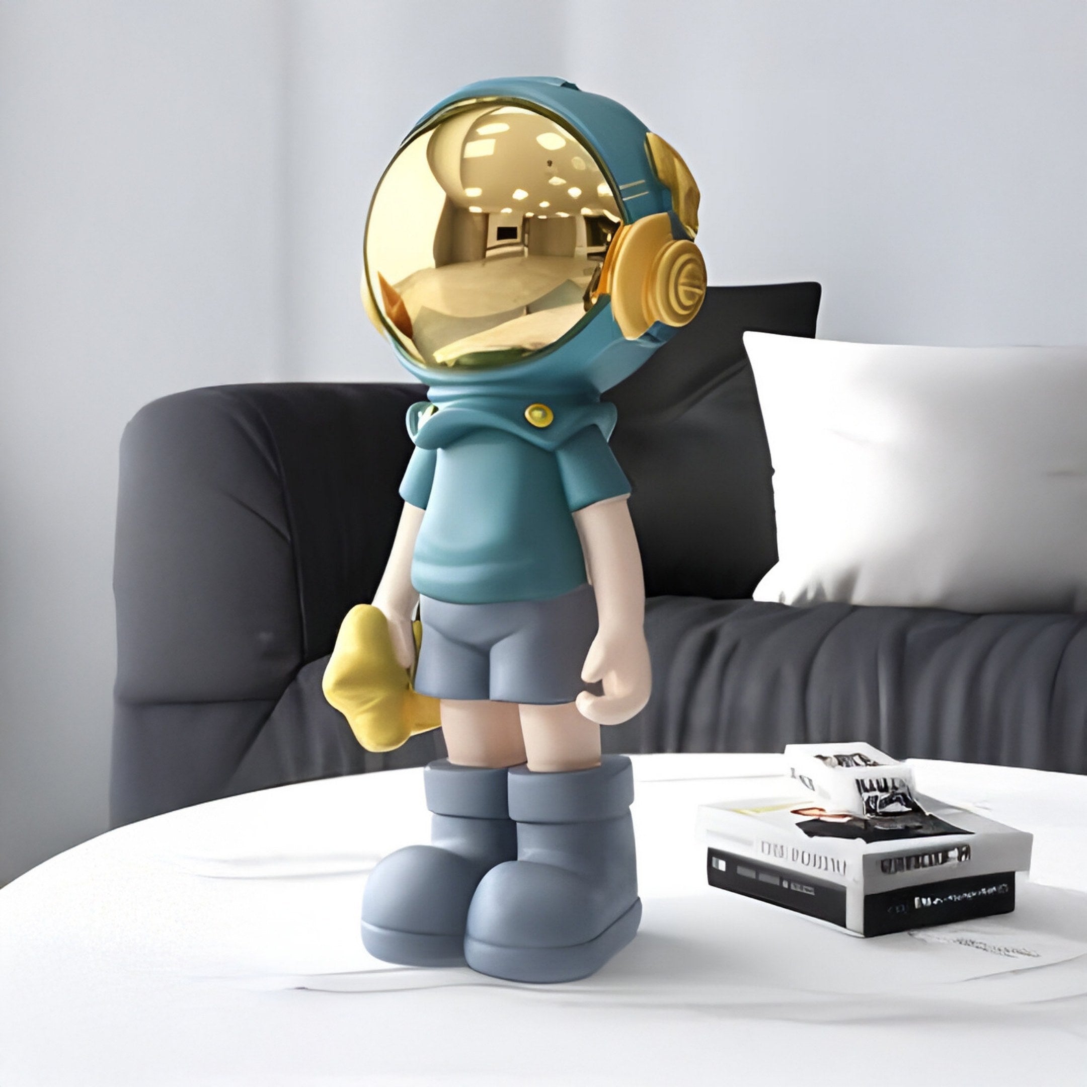 This Galactic Blue Astronaut Buddy Figure from Giant Sculptures features a gold helmet and vibrant blue outfit. It holds a yellow object, positioned on a white table in a modern living room with a black sofa and books.