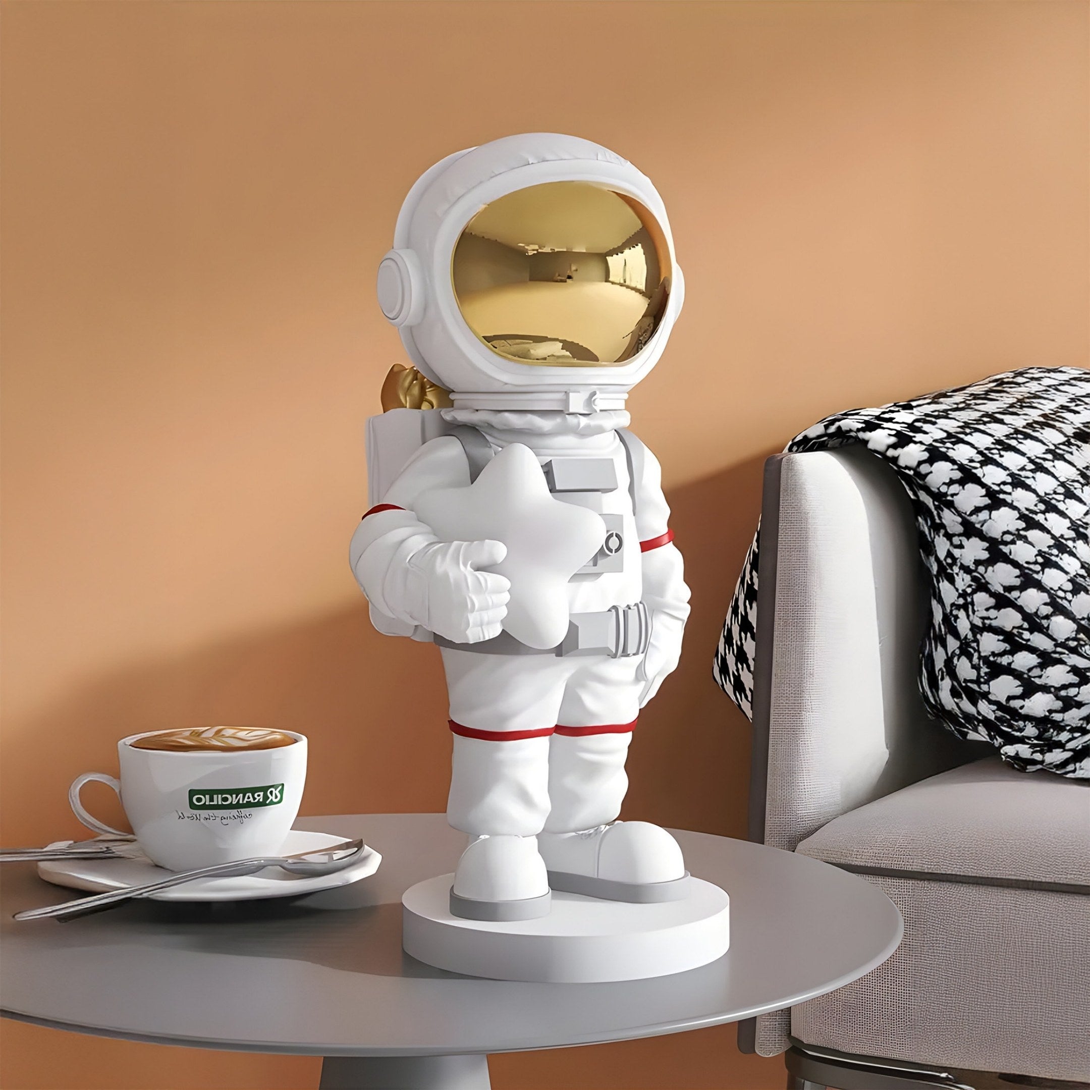 A Giant Sculptures Nova White Astronaut Star Figure with a shiny gold visor stands on a round gray table, adding imaginative home accents. Nearby, a coffee cup highlights latte art, and a houndstooth-patterned blanket is draped over a chair in the background.