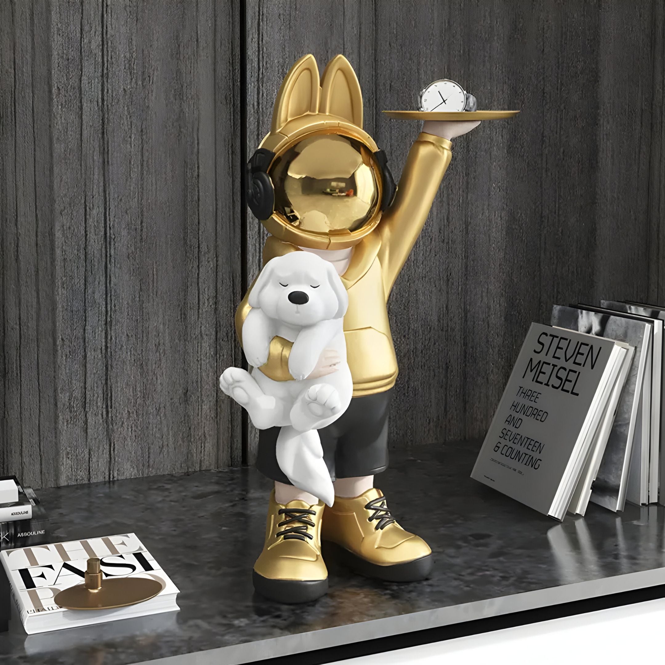 The Gold Astronaut Bunny Pup Butler Figure - 44cm by Giant Sculptures features a golden, bunny-eared statue with headphones holding a white dog and clock. Its displayed on a shelf with books, like STEVEN MEISEL, set against a wooden backdrop.