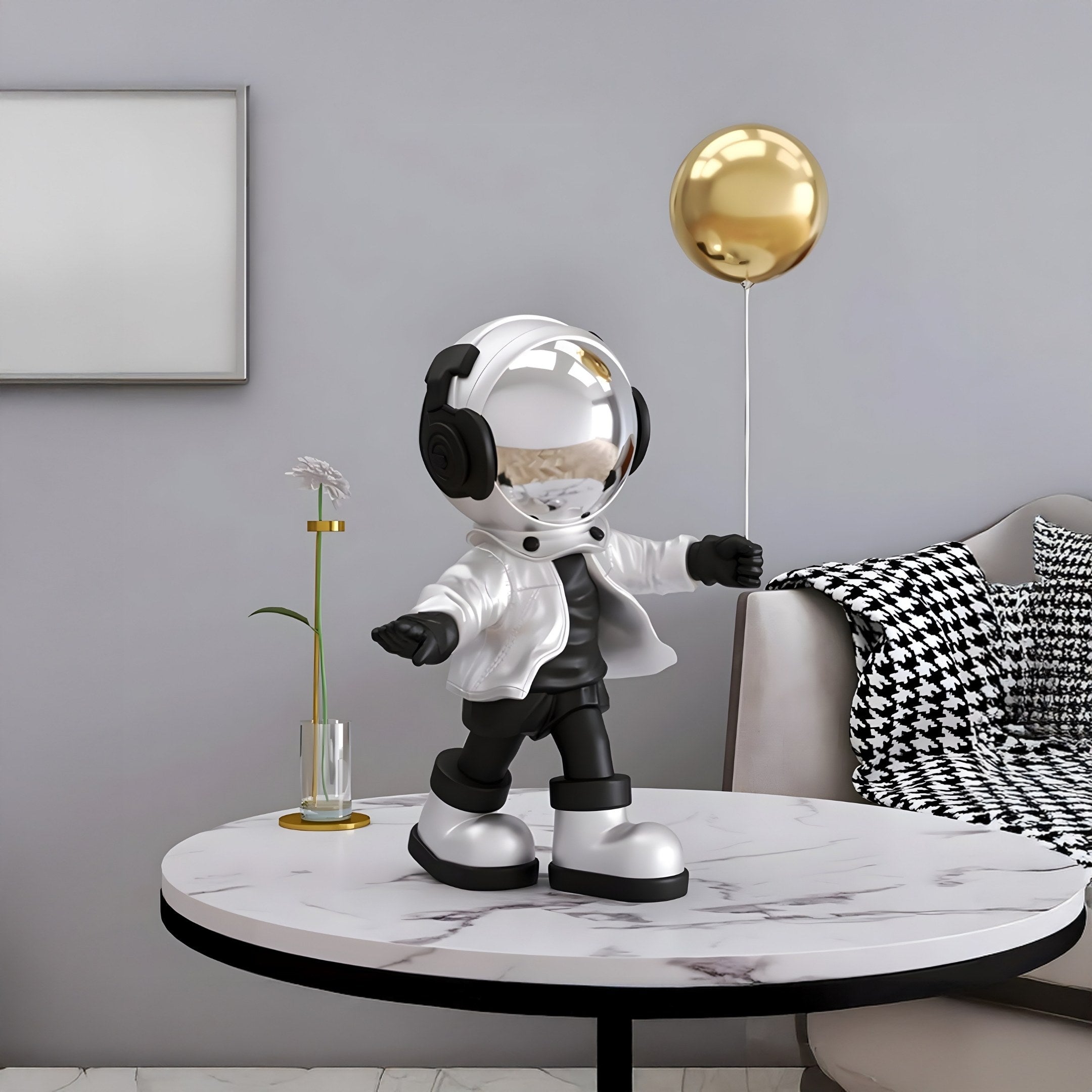 A 48cm Lunar Silver Astronaut Balloon Standing Figure by Giant Sculptures stands on a marble table with a gold balloon, its gold helmet gleaming. A vase with a white flower and a couch with black-and-white cushions create contrast against the gray wall for a futuristic decor vibe.