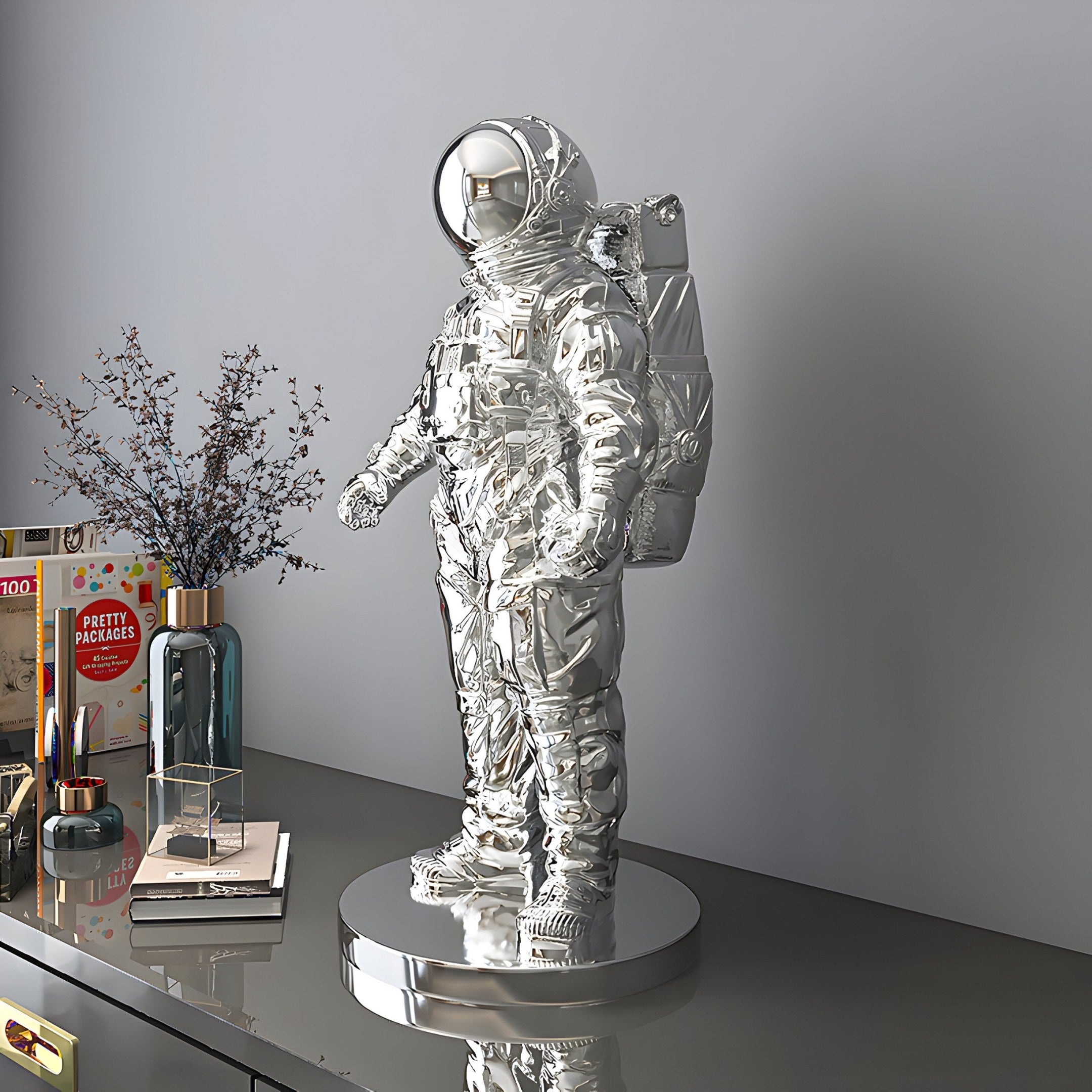 The Giant Sculptures Cosmic Chrome Space Astronaut Standing Sculpture - 58cm with a reflective finish stands on a metallic base atop a modern gray cabinet surrounded by decorative items—a vase with branches, a candle, and books—bathed in soft lighting.