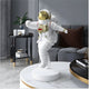 The White Space Astronaut Strider Sculpture - 58cm by Giant Sculptures, with a reflective gold helmet, stands on a round white base on a marble table. A gray wall, decorated side table, and dark sofa with brown cushions enhance the elegant cosmic ambiance.