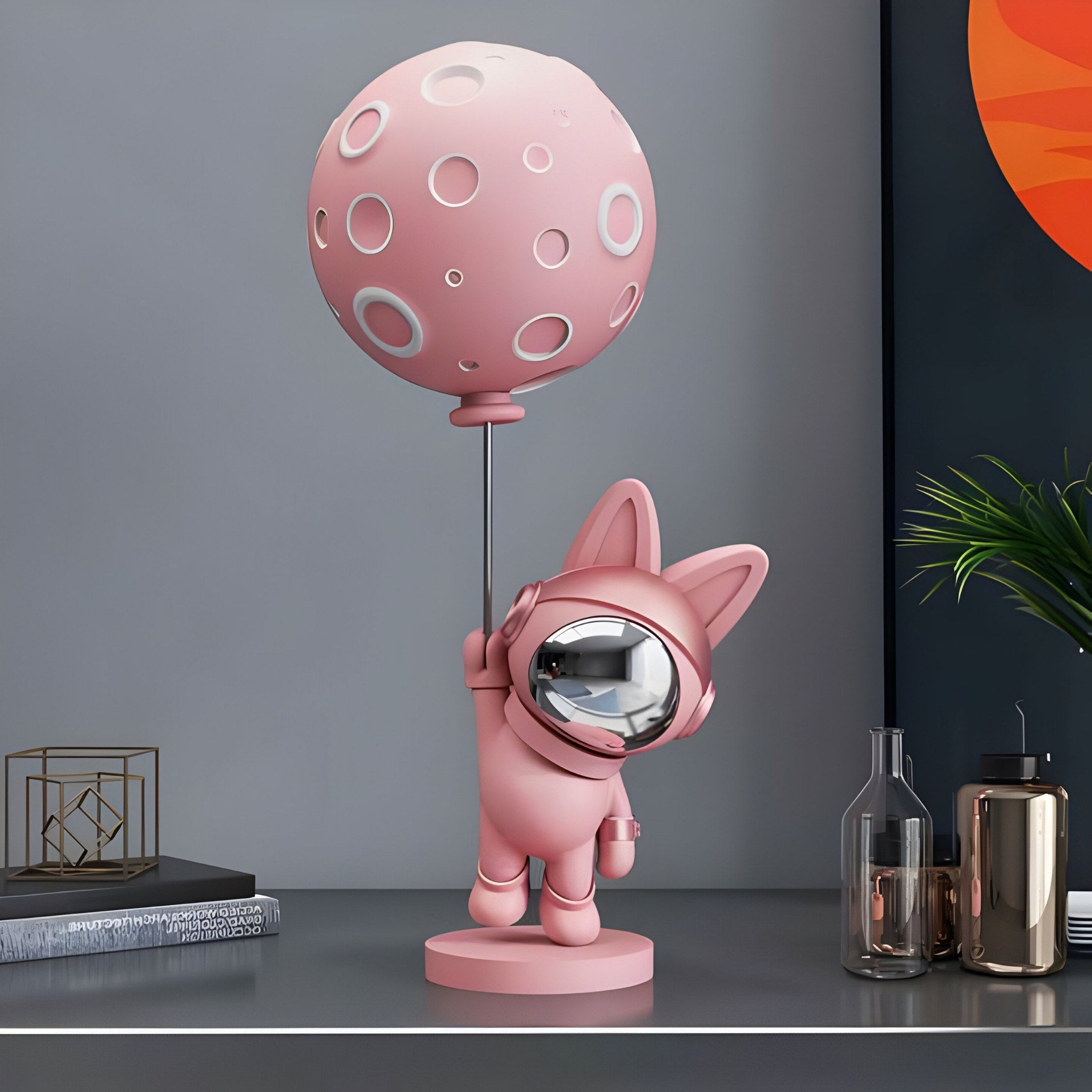 The Candy Pink Astronaut Bunny Moon Balloon Sculpture (60cm) by Giant Sculptures features a bunny holding a rod with a round, perforated moon balloon. Its shiny helmet gleams among décor, adding whimsy and charm to any space on a gray surface with decorative books, bottles, and plants nearby.