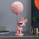 The Candy Pink Astronaut Bunny Moon Balloon Sculpture (60cm) by Giant Sculptures features a bunny holding a rod with a round, perforated moon balloon. Its shiny helmet gleams among décor, adding whimsy and charm to any space on a gray surface with decorative books, bottles, and plants nearby.