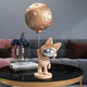 The Rose Gold Astronaut Bunny Moon Balloon Sculpture from Giant Sculptures, with its reflective visor and rabbit ears grasping a balloon pole, sits on a coffee table with books and a glass vase of blue flowers. It complements the gray and pink tones of the sofa beautifully.