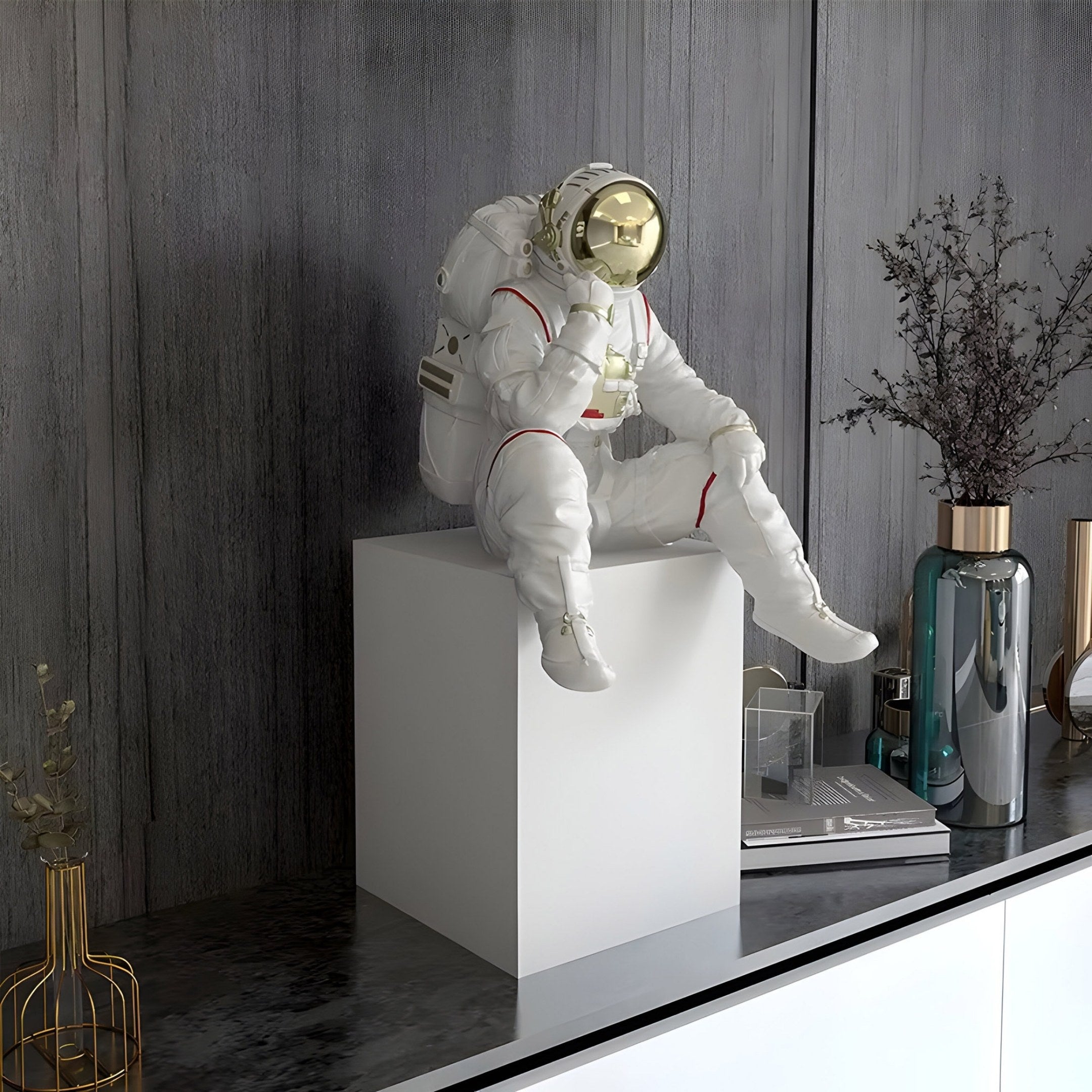 A 65cm Nova White Astronaut Thinker Sculpture by Giant Sculptures, sporting a reflective gold helmet, sits elegantly on a white pedestal atop a modern dark-toned shelf with space-themed decor. The setting includes vases, books, and a textured gray wall backdrop.