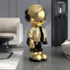 The Celestial Gold Astronaut Beats Sculpture by Giant Sculptures, with its reflective gold and black accents, stands elegantly in a modern minimalist room. It contrasts beautifully against gray walls, a white armchair, and a contemporary lamp, perfectly complementing the space-themed decor.