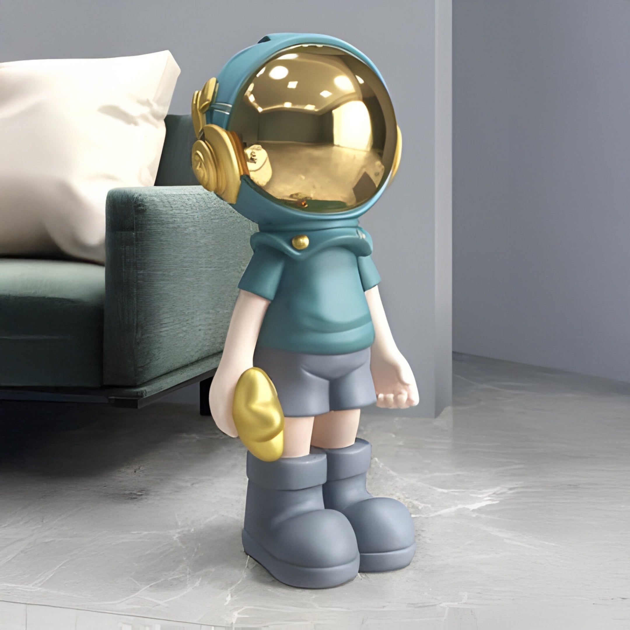 The Galactic Blue Astronaut Buddy Sculpture by Giant Sculptures, standing at 70cm, features a glossy gold helmet and blue outfit with boots. Holding a gold shape, it stands on a marble floor, ideally complementing the teal sofa in the space-themed décor setting.