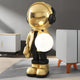 The Celestial Gold Astronaut Glow Orb Sculpture - 80cm by Giant Sculptures, features a poised gold astronaut with a large helmet and headphones holding a glowing white orb. This statement piece illuminates the modern floor, complementing the light gray walls and white-gray furniture around it.