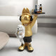 The Gold Astronaut Bunny Pup Butler Sculpture by Giant Sculptures features a bunny with headphones and animal ears, cradling a white puppy and balancing a tray of bottles. At 80cm tall, it adds whimsical flair as a centerpiece near a white couch.