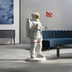 The White Space Astronaut Pioneer Standing Sculpture - 80 cm by Giant Sculptures adds a futuristic touch to a modern living room, featuring a white suit and gold visor. Paired with a red flag, it complements space enthusiast decor including a blue sofa, black sideboard, and abstract wall art.