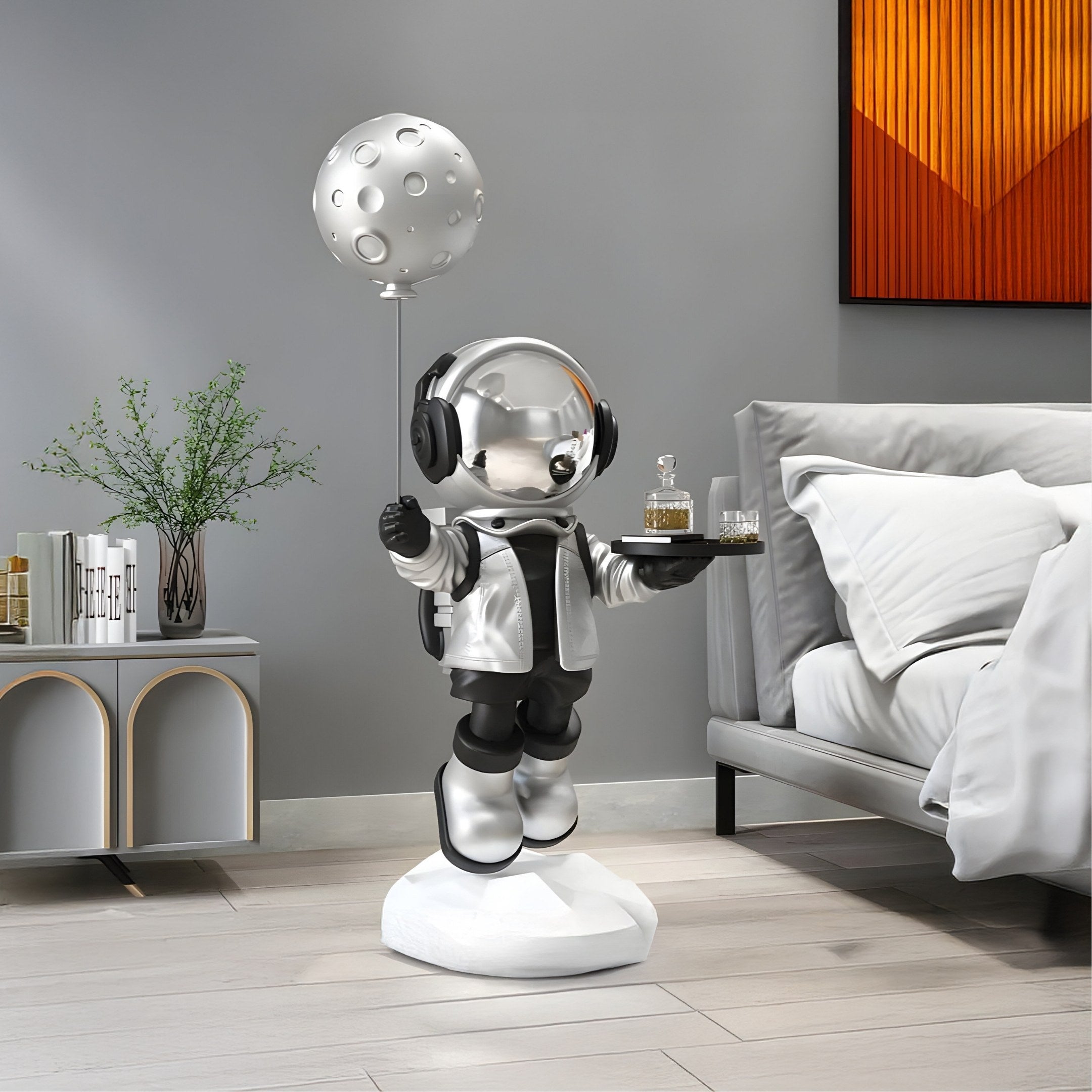 The Lunar Silver Astronaut Butler Balloon Sculpture (80cm) by Giant Sculptures stands on a white base holding a moon balloon and drink tray, enhancing a modern bedroom with its cosmic charm alongside plants, a small table, and an orange abstract painting.