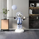 In a contemporary room stands the Cosmic Sky Astronaut Butler Balloon Sculpture by Giant Sculptures, showcasing an astronaut holding a moon-patterned balloon and tray with a moon model. The backdrop includes a beige wall, wooden side table with plant, and bookshelves filled with indoor decor items.