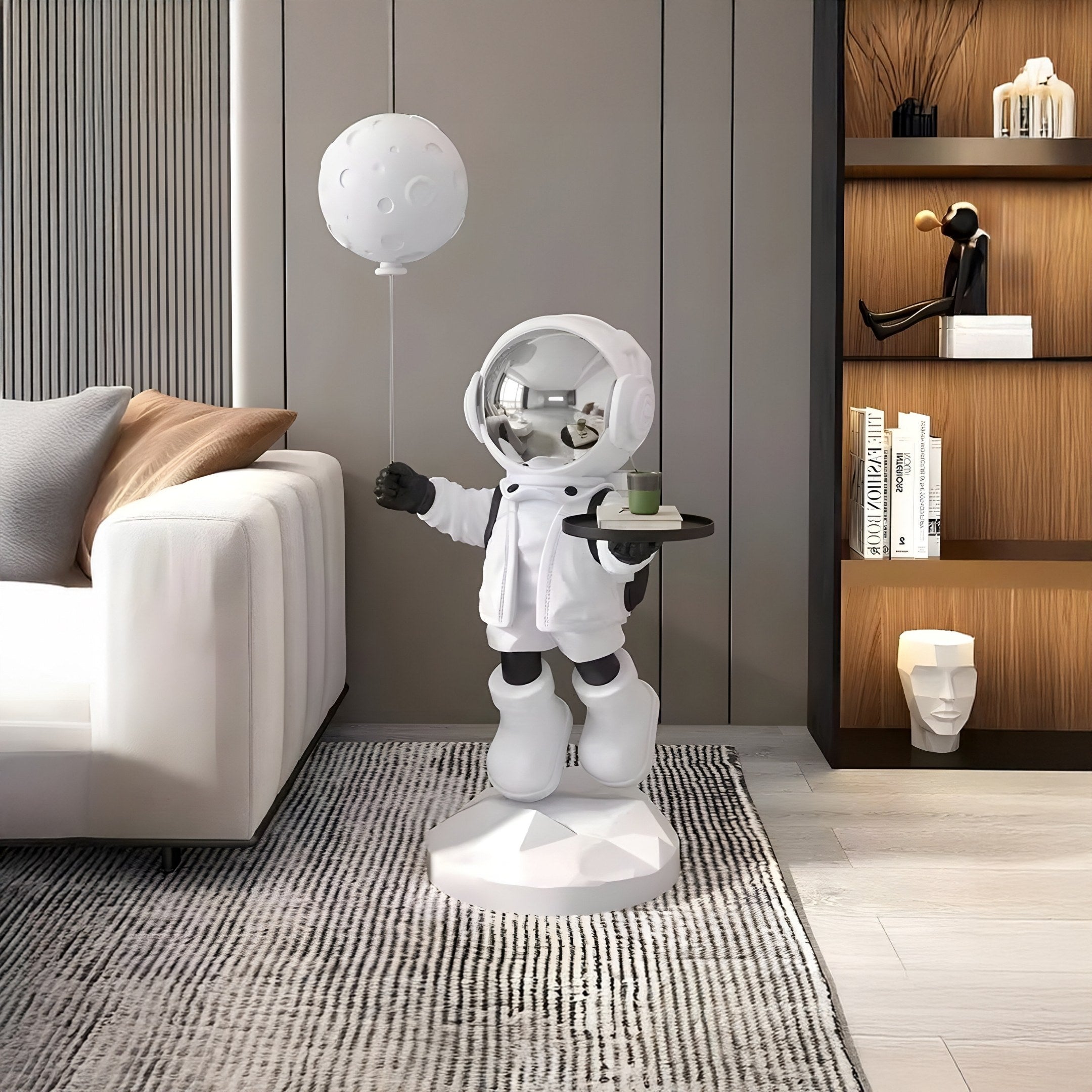 The Nova White Astronaut Butler Balloon Sculpture by Giant Sculptures adds cosmic flair with its tray and balloon design. Positioned on a geometric base, it complements the modern gray-cushioned couch as wooden shelves display books and abstract decor in the background.