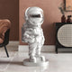 The 85cm Lunar Silver Astronaut Star Sculpture by Giant Sculptures stands on a round base, shining brilliantly against the warm brown walls of the modern room, complemented by a chair and sideboard with decorative items in the background.