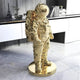 The Gilded Gold Space Astronaut Standing Sculpture by Giant Sculptures gleams on a round base atop the marble floor, with white cabinets behind adorned with a small Eiffel Tower model and books, creating a futuristic home décor touch.