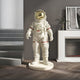 In a sleek, minimalist space, the Cloud White Space Astronaut Standing Sculpture by Giant Sculptures (88cm) captivates with its reflective gold helmet on a pedestal. The design features a left-side staircase and a tastefully decorated white fireplace on the right.