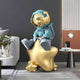 A Galactic Blue Astronaut Beats On Star Sculpture by Giant Sculptures, 90cm tall with a gold visor, complements the modern living rooms white sofa, lush plants, and sleek side table with a lamp to create a serene atmosphere.