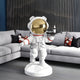 The Nova White Astronaut Bear Butler Sculpture - 90cm by Giant Sculptures features a gold-helmeted bear holding a tray with a bottle. Positioned on its white base, it enhances the imaginative decor of a modern living room, complementing the gray sofa and geometric cushions against striped wall panels.