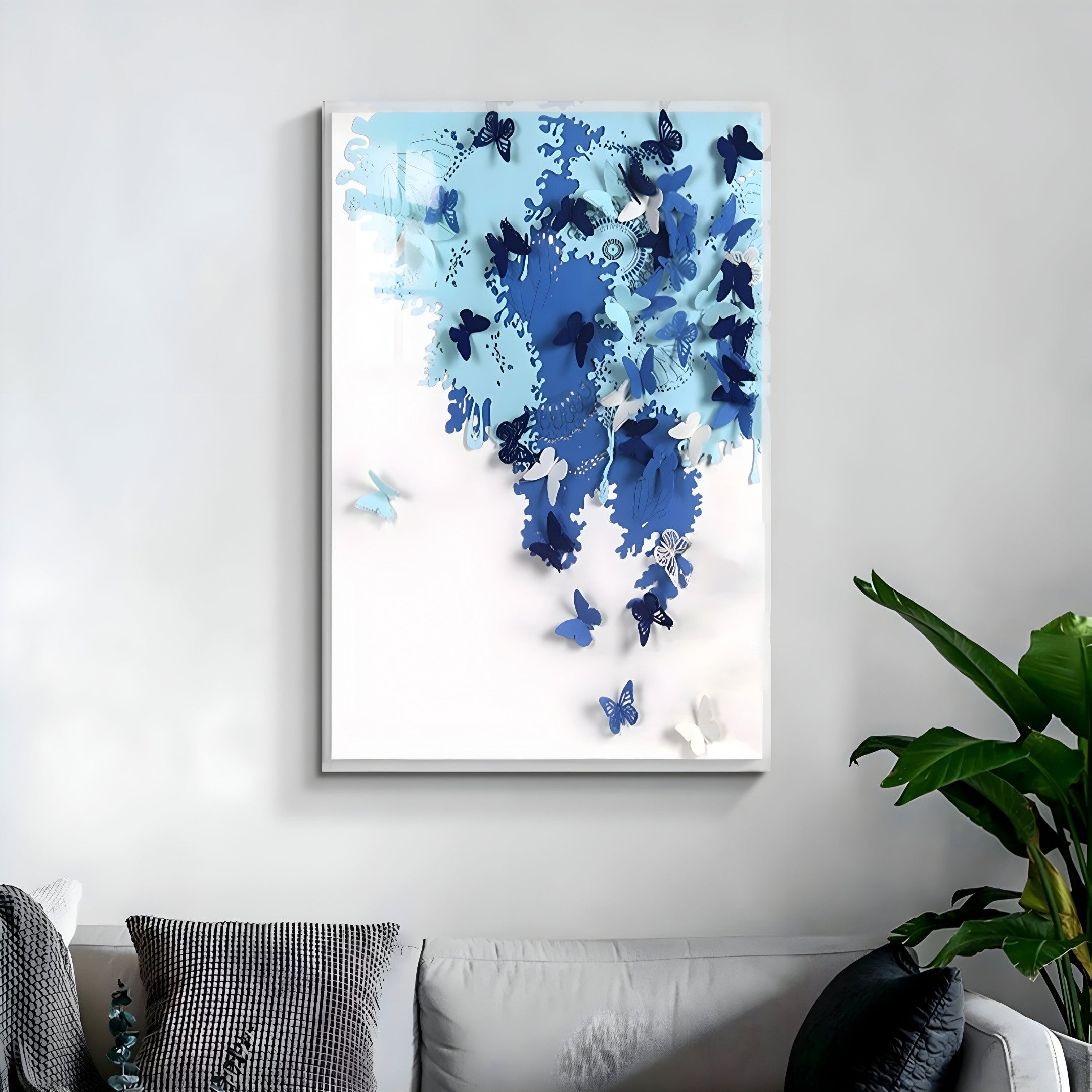 A wall features the Luminara Blue II Butterfly Paper 3D Wall Art by Giant Sculptures, showcasing blue and white butterflies in a gradient, densely clustered and dispersing. Below, a gray sofa with monochrome cushions sits beside a lush green plant.