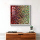 The Chromatica Square Radiant Red & Green Rolled Paper 3D Wall Art by Giant Sculptures, showcasing a vibrant mosaic of varied hues, adorns a white wall. Below it, a wooden cabinet displays a small potted plant and stacked magazines.