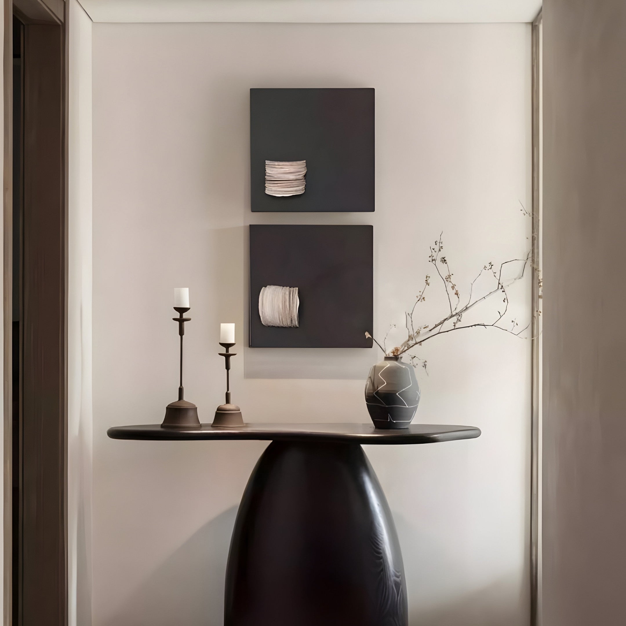 A minimalist hallway showcases a round black table with a vase and twig, while the Modulus Black & White Duo Panel Abstract Wall Art by Giant Sculptures hangs above. Tall candlesticks with white candles adorn the table, enhancing the harmonious ambiance.