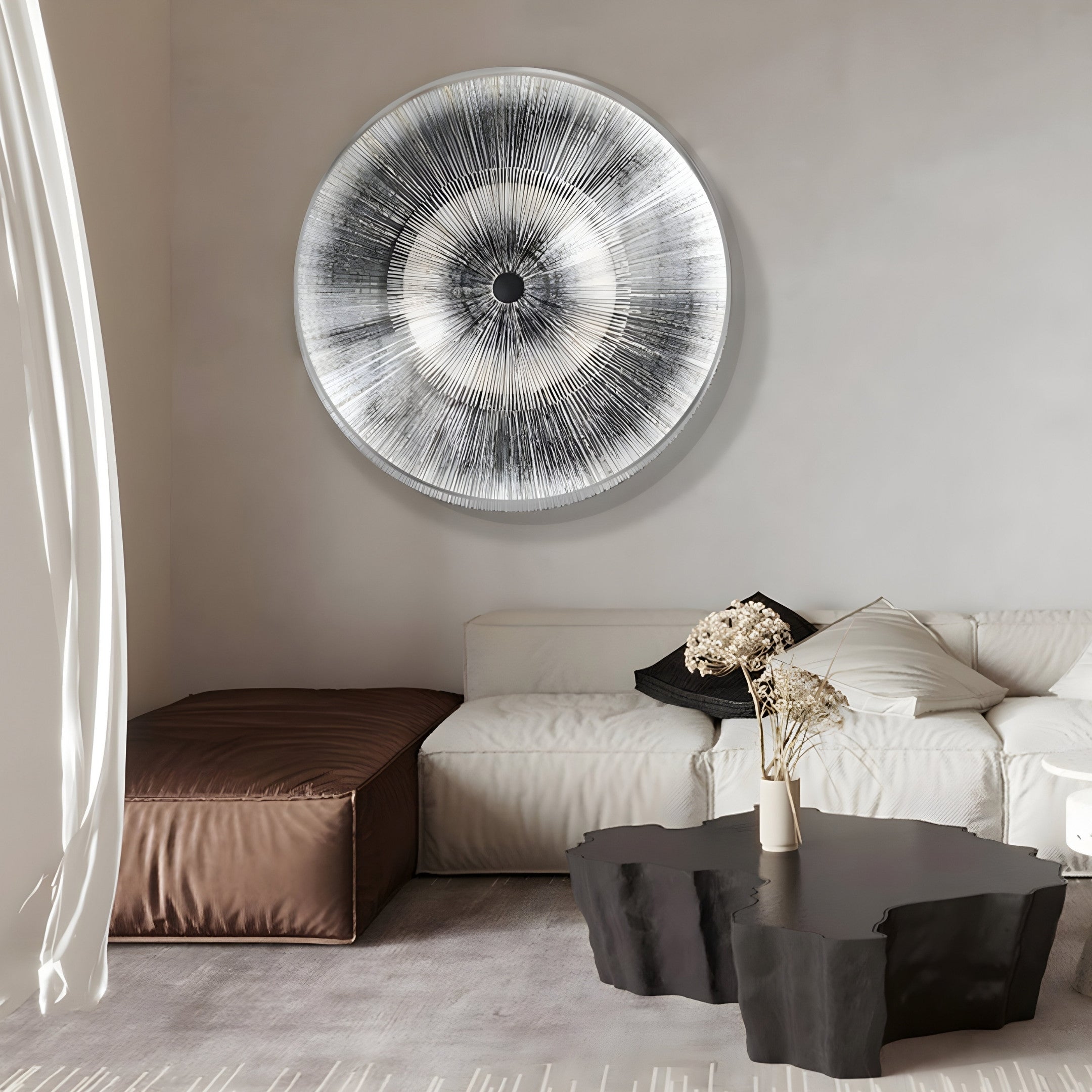 A modern living room showcases the Oculis Monochrome Circular Handmade 3D Paper Wall Art by Giant Sculptures above a white sectional sofa. A brown ottoman sits next to it, while a black coffee table holds a vase of dried flowers. White curtains nearby enhance the clean, contemporary vibe.